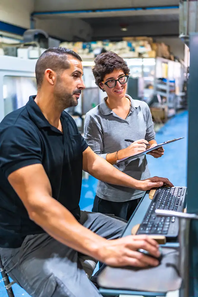 Engineer at computer workstation reviewing data with colleague for purchasing reports